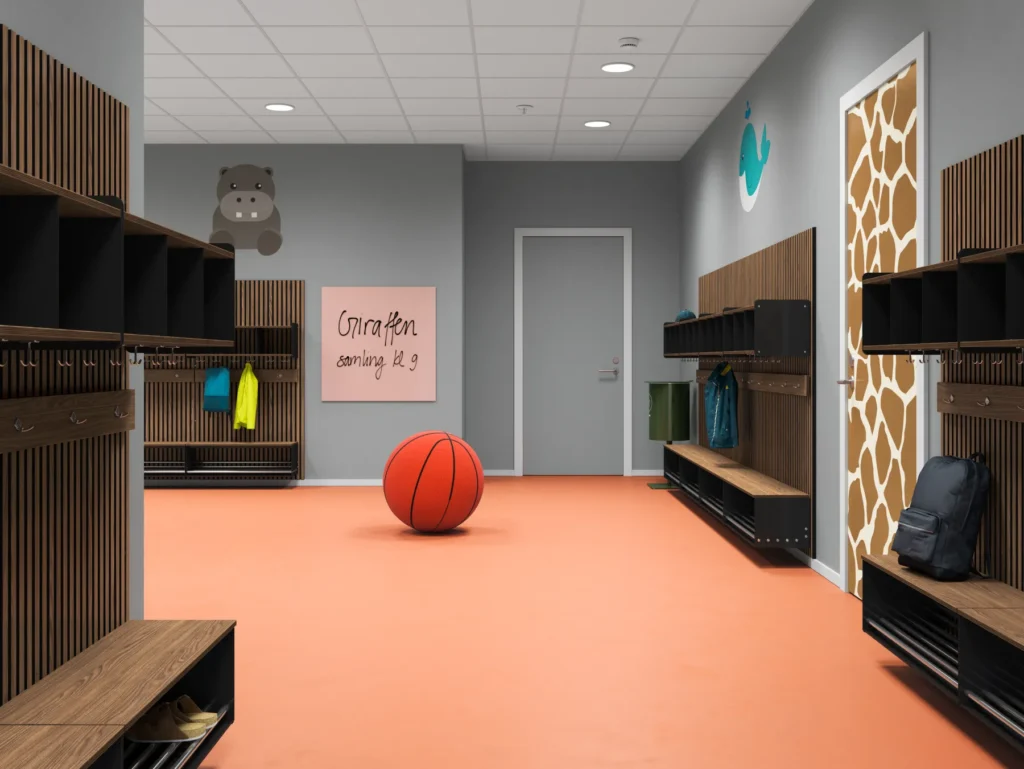 Förskolehall med orange golv, grå väggar och väggmålningar av en blå val och en flodhäst. Inredningen är i ljus trä med grå metallramar – med hyllor, krokar och sittbänkar längs båda sidor. En dörr med giraffmönstrad dekor står till höger, och i mitten av rummet ligger en stor leksaksbasketboll. En rosa skylt visar texten 'Giraffen samling kl 9'.