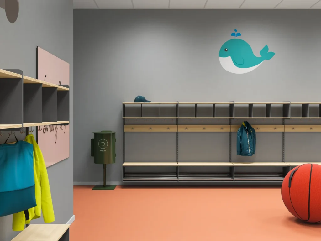 Förskolehall med orange golv och grå väggar, dekorerad med en blå val. Inredningen består av ljusa träbänkar och hyllor med krokar undertill. En jacka och keps hänger vid facken. Till vänster syns klädhängare och ett rosa konstverk, och på golvet står en grön soptunna för organiskt avfall samt en stor leksaksbasketboll.