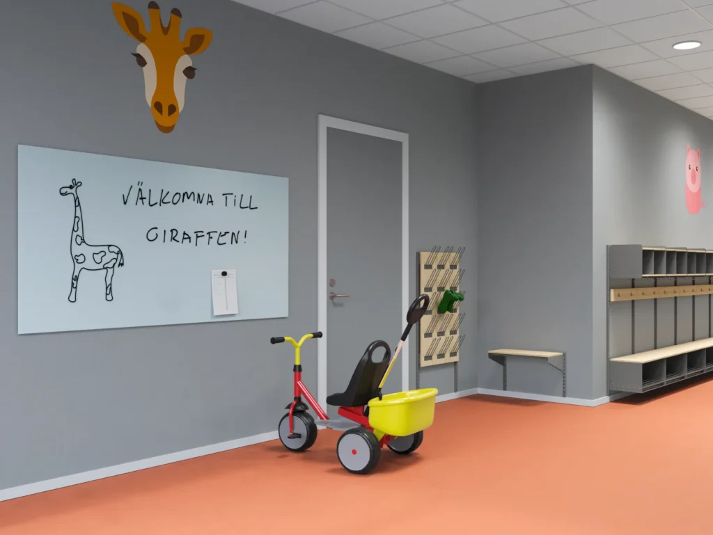 Förskoleentré med orange golv, grå väggar och ett stort giraffhuvud som väggmålning. En whiteboard visar texten 'Välkomna till Giraffen!' tillsammans med en tecknad giraff. En trehjuling med gul skopa står vid dörren. På högra väggen finns ljusa förvaringslösningar med krokar och sittbänk, samt ett skoställ i trä och metall. En rosa gris är målad på väggen längre bort.
